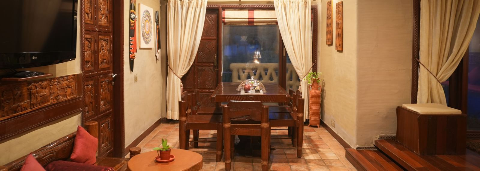 Living area of the Zamindar Suite in Vedic Village Spa Resort featuring an in-room dining space, wooden furniture, ambient lighting, and a flat-screen TV