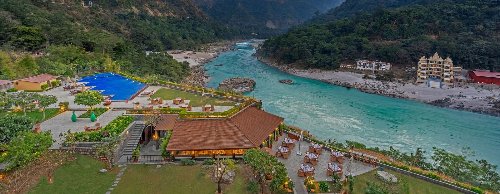 A panoramic view of Aloha on the Ganges, Rishikesh. The image shows the resort nestled amidst hills, with the Ganges River flowing peacefully in the foreground. There is a swimming pool, restaurant, and other facilities visible.