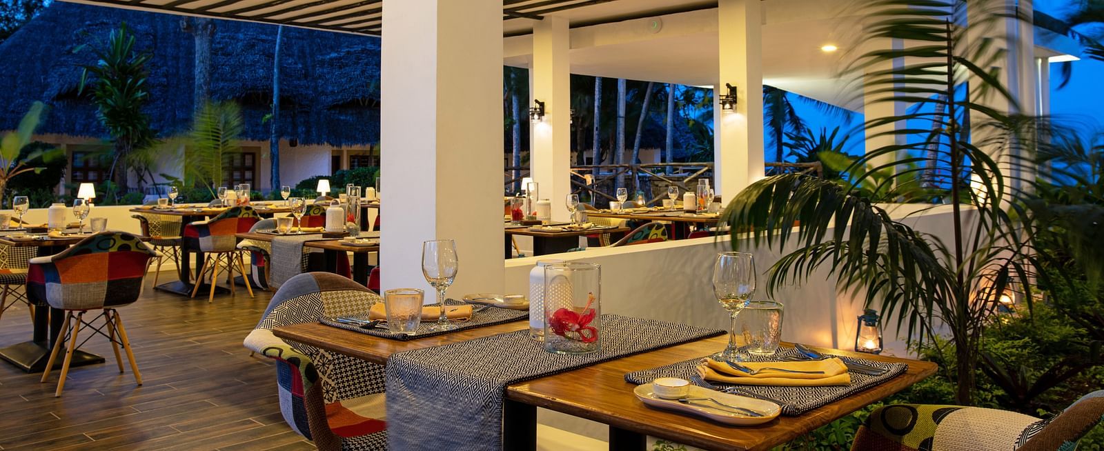 A well-lit indoor restaurant dining area with wooden tables set with glasses, plates and napkins, surrounded by patterned chairs and decorative lighting at shooting star Boutique Hotel
