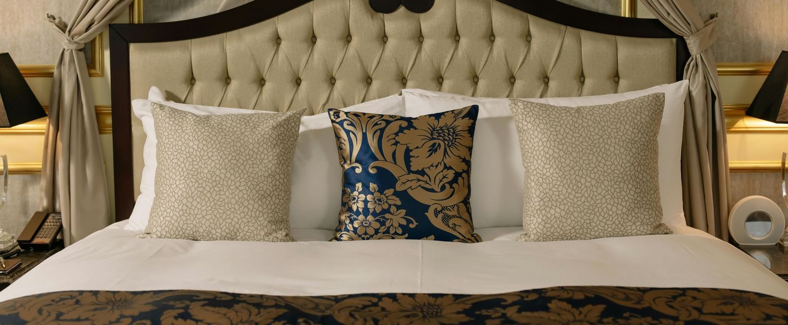 A luxurious hotel room features a large bed with a canopy and elegant bedding, flanked by bedside tables with lamps.