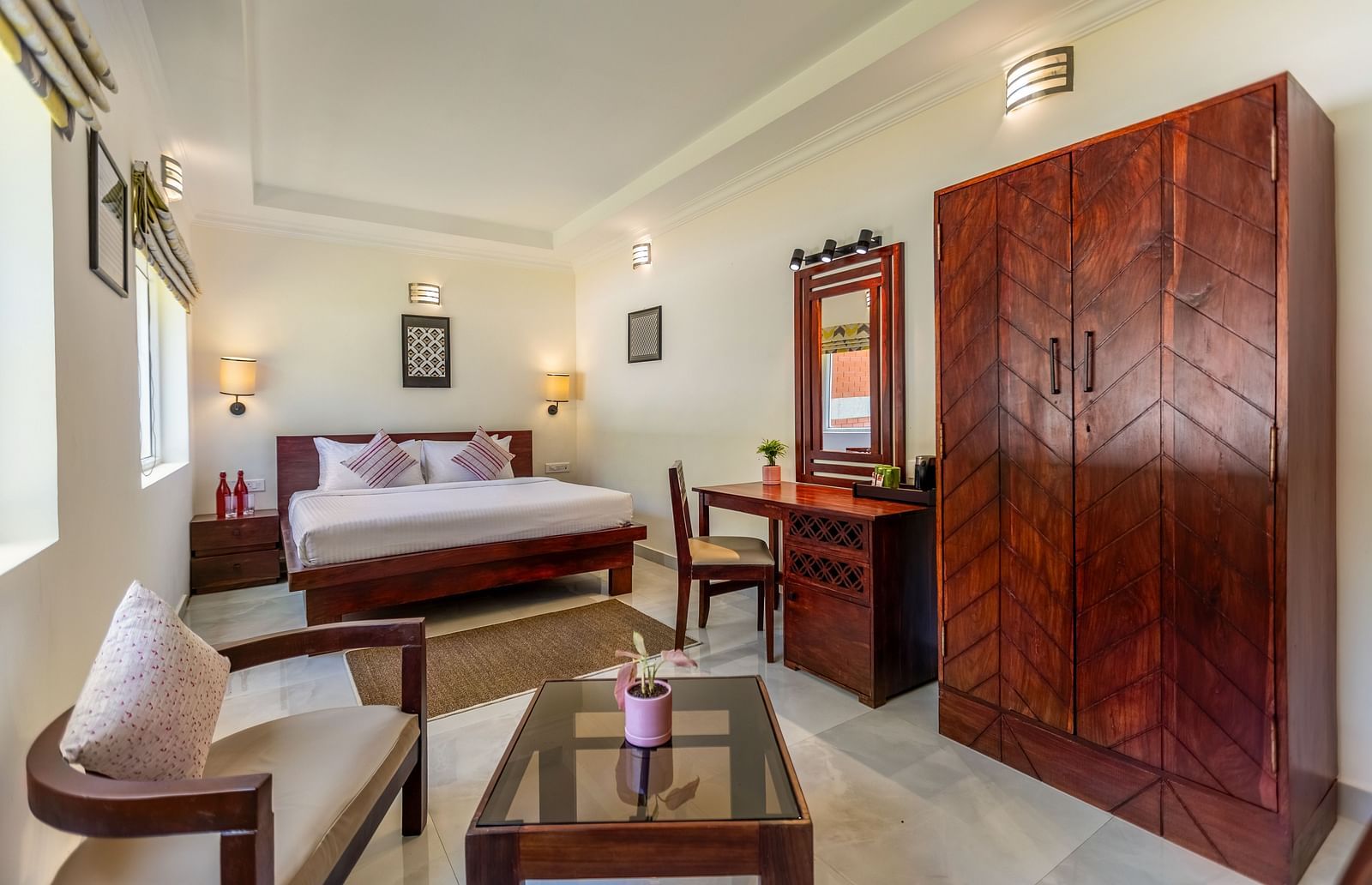 A bright, clean hotel room with a bed, a wooden desk and chair, a large wooden wardrobe, and a seating area with a glass-topped coffee table - Mango Hill Resort, Coimbatore