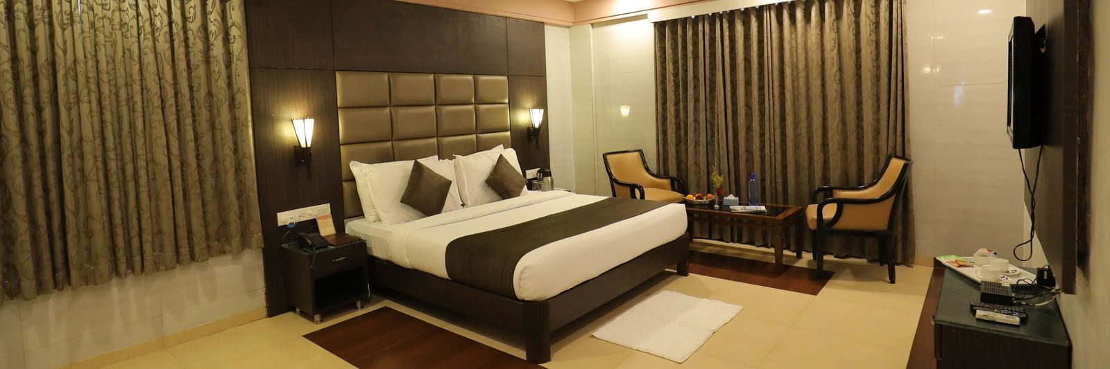 A front angle view of a bedroom at Krushnai Resort with a king size bed, a dark headboard, and brown drapes.