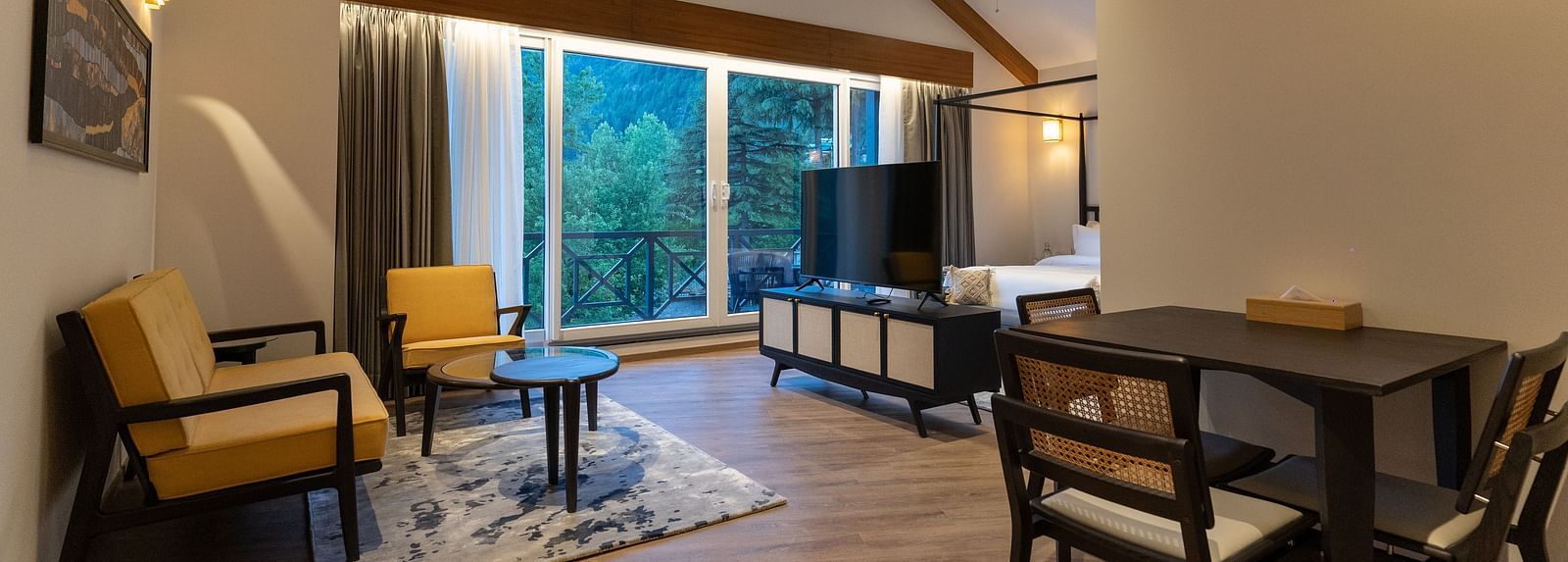 Spacious room with cushioned sofa, wooden table with a TV, and large windows offering scenic nature views at Nivasana Manali.