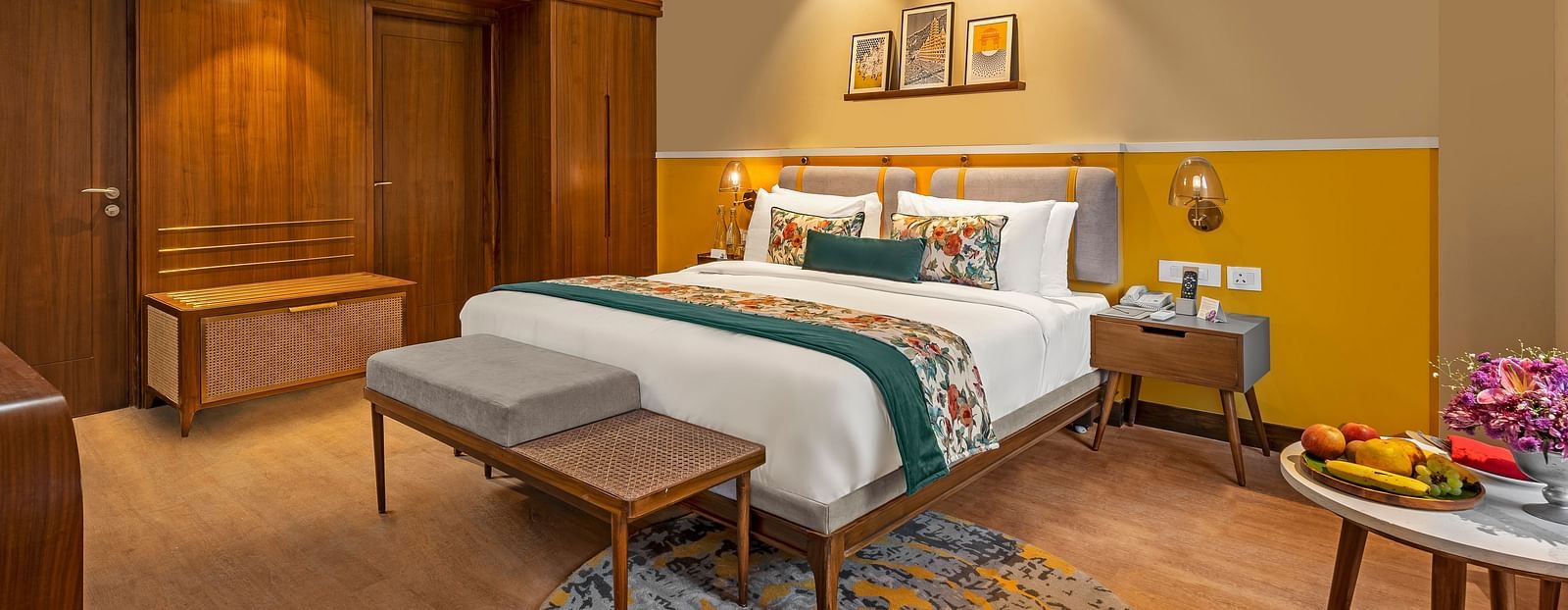 An image of a room featuring a king-size bed with a headboard and comfortable bedding. A bench sits at the foot of the bed, and a small table with a fruit platter is placed nearby.