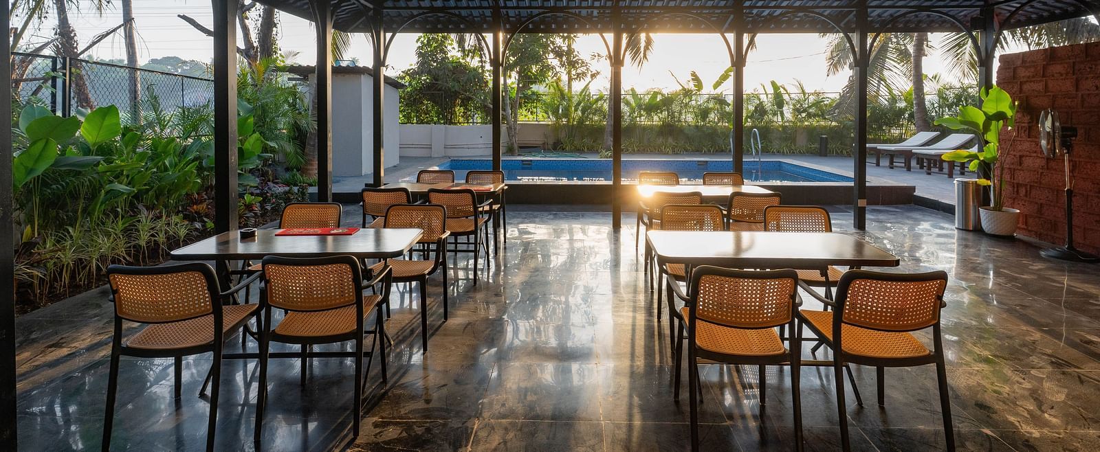 An outdoor dining area under a dark, peaked roof, open to the surrounding scenery. Several wooden tables and chairs are neatly arranged, with a glimpse of a swimming pool visible in the background under the bright sky at Kssemya