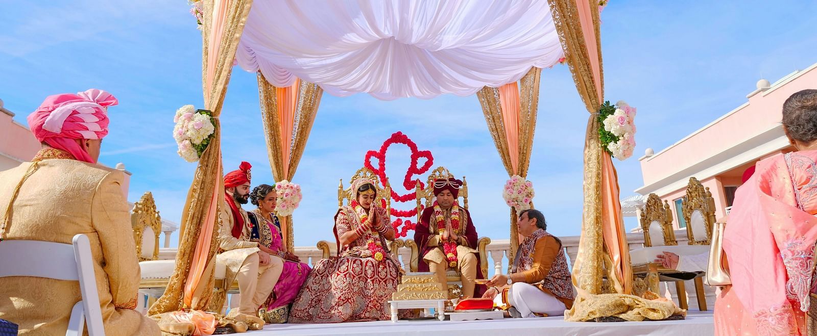 A wide angle shot of an Indian wedding happening under a gazebo decorated with beautiful flowers and drapes with a clear blue sky in the background. 