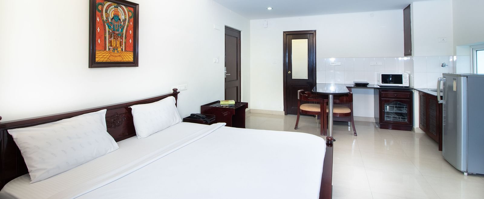A room with a double bed featuring white linens, a ceiling fan, a framed picture on the wall, a dining counter, a refrigerator, and the kitchenette area at Sanctum Suites Indiranagar, Bangalore.