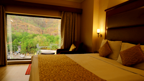 A neat double bed in a warmly lit Hill View Room featuring a huge glass window with scenic mountain views at Raj Park Hotel, Tirupati.