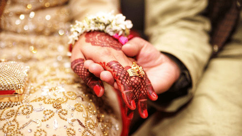 Close-up of two people holding hands; one hand has henna, red nails, gold rings, and a flower bracelet, resting on ornate gold-embroidered fabric, suggesting a wedding setting.