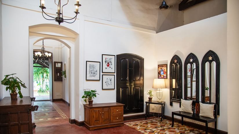 An image of a huge lobby where various decorative items are placed on top of the cabinets and shelves, there is another table with lamp and three vintage style window near it - Lamrin Ucassaim Hotel, Goa