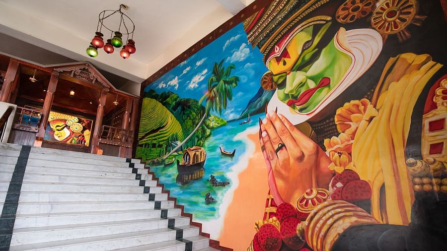 A vibrant mural featuring a traditional Kathakali dancer, tropical scenery, and boats decorates the wall beside a wide staircase at Jenneys Residency with ornate wooden carvings and a hanging chandelier