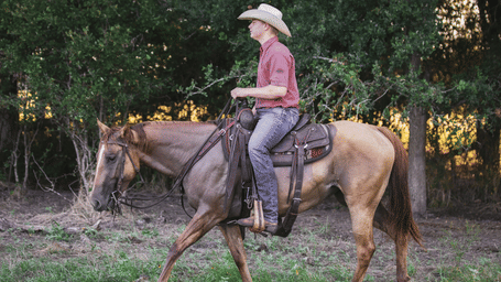 Side view of a person in a cowboy hat and jeans riding a light brown horse through a grassy field near woods at sunset.
