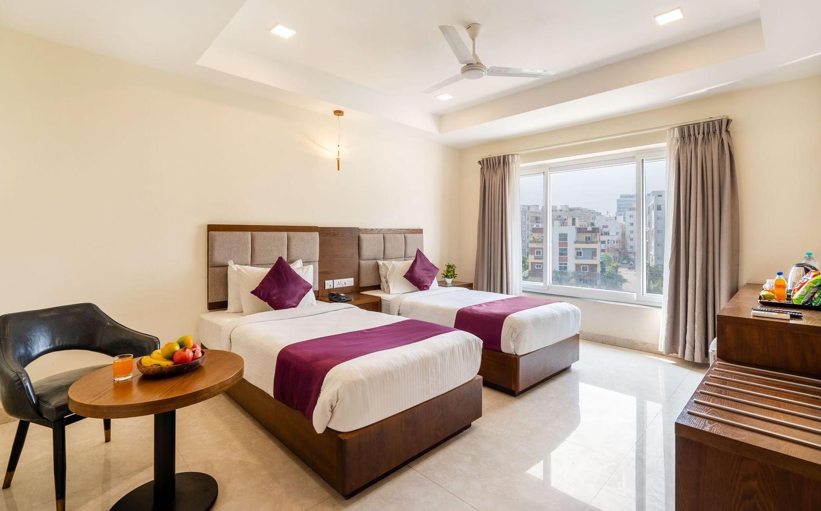 A Deluxe Room at Destino by Monday Hotels, Gachibowli, with twin beds, a small table with fruits, and a large window providing natural light.