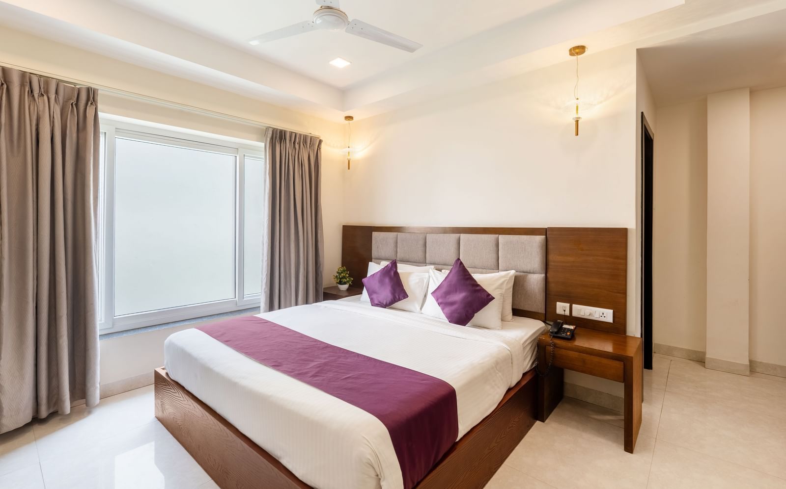 A Premium Room at Destino by Monday Hotels, Gachibowli, with a large double bed with purple accents and a ceiling fan, illuminated by natural light from a large window.