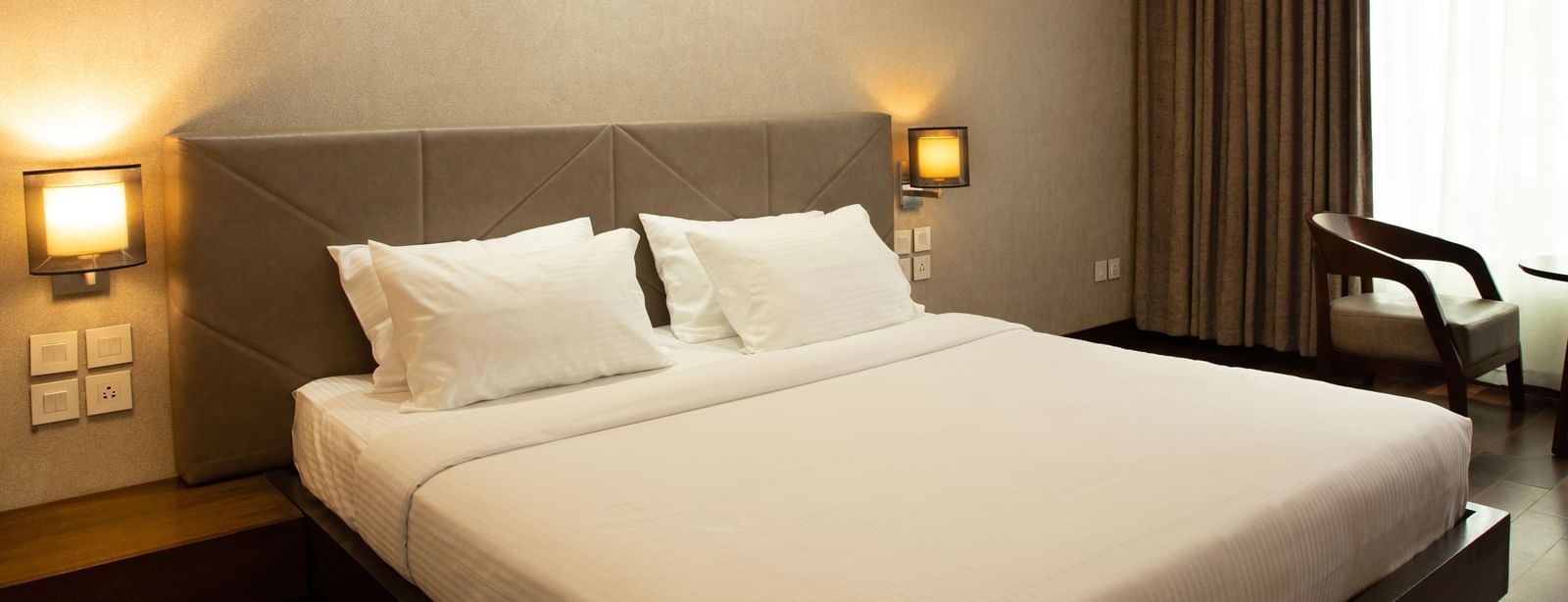 A bedroom with a large bed, bedside lamps, curtains drawn to the side, and natural light coming through the window at The Hamuse Hotel, Kodaikanal.