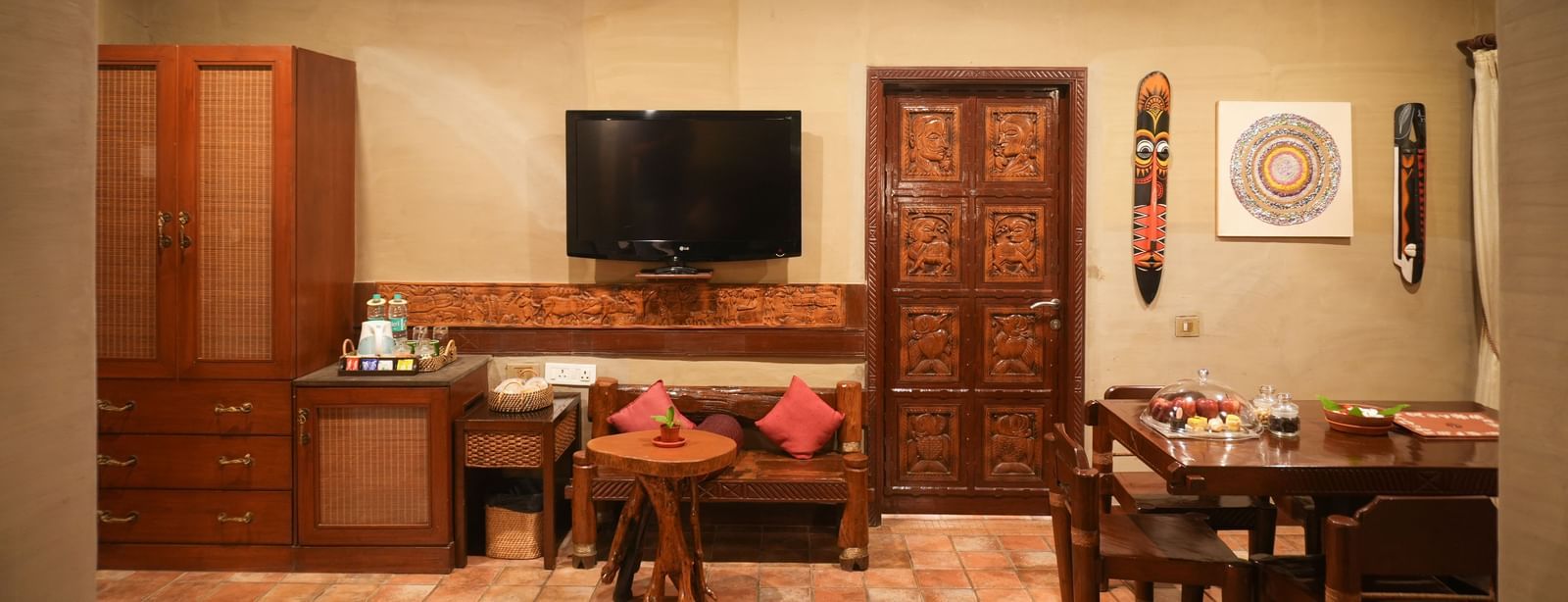 Living area of the Zamindar Suite at Vedic Village Spa Resort with an in-room dining setup, warm ambient lighting, wooden furnishings, and a wall-mounted TV.
