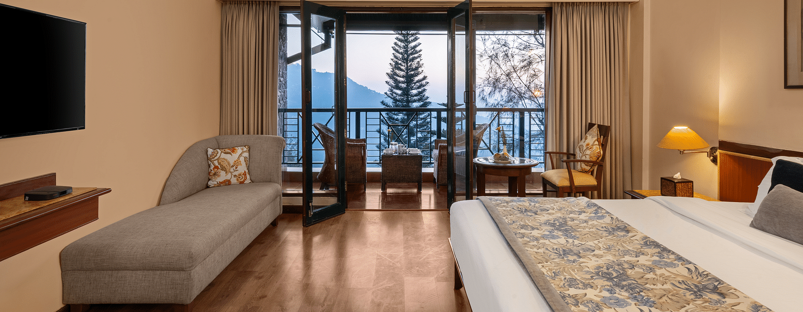 A superior room with a neatly made bed, a TV mounted on the wall, a side sofa, and a balcony featuring a wooden chair and table -  Baikunth resort, kasauli 
