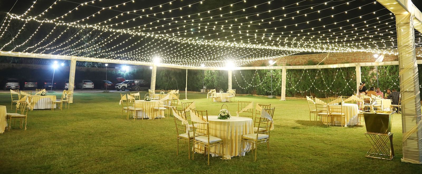 A large lawn at Tisya Farms, Gurgaon with round tables, chairs, and string lights set up for an evening event.
