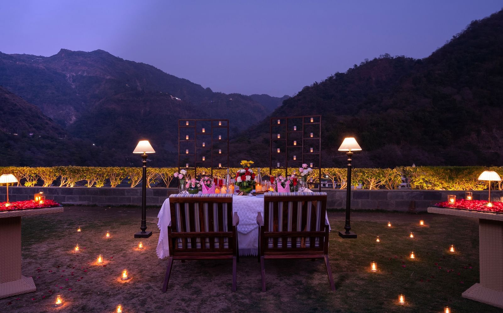 A dinner setup outdoors with a table adorned with candles and flowers,he backdrop is a beautiful mountain range at dusk.