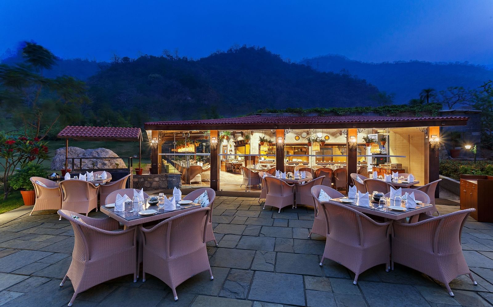An outdoor restaurant with a view of mountains at dusk. Tables and chairs are set up, a covered area with warm lighting in the background.