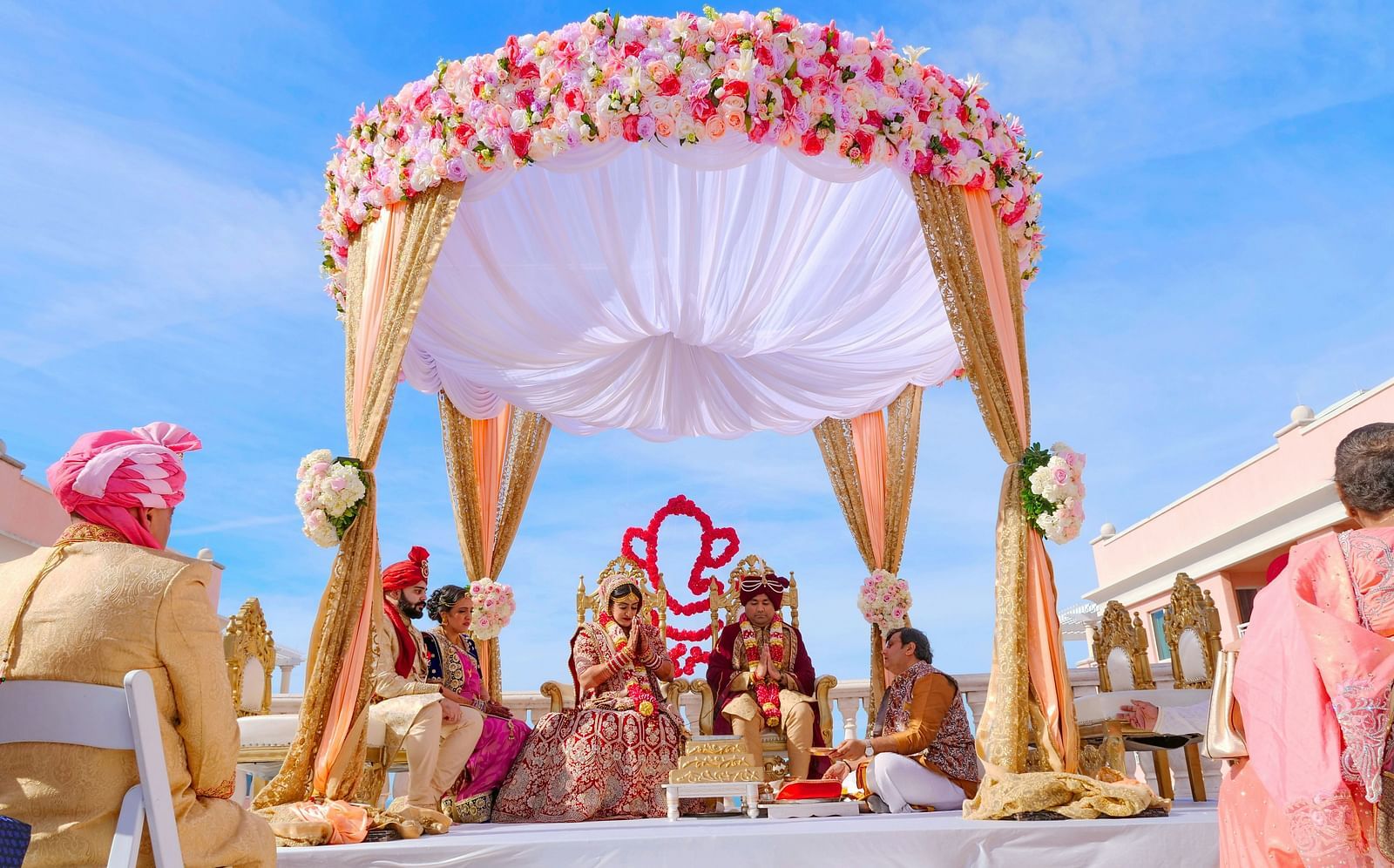 This image showcases a grand Indian wedding ceremony taking place outdoors under a beautiful, floral-adorned setup. 