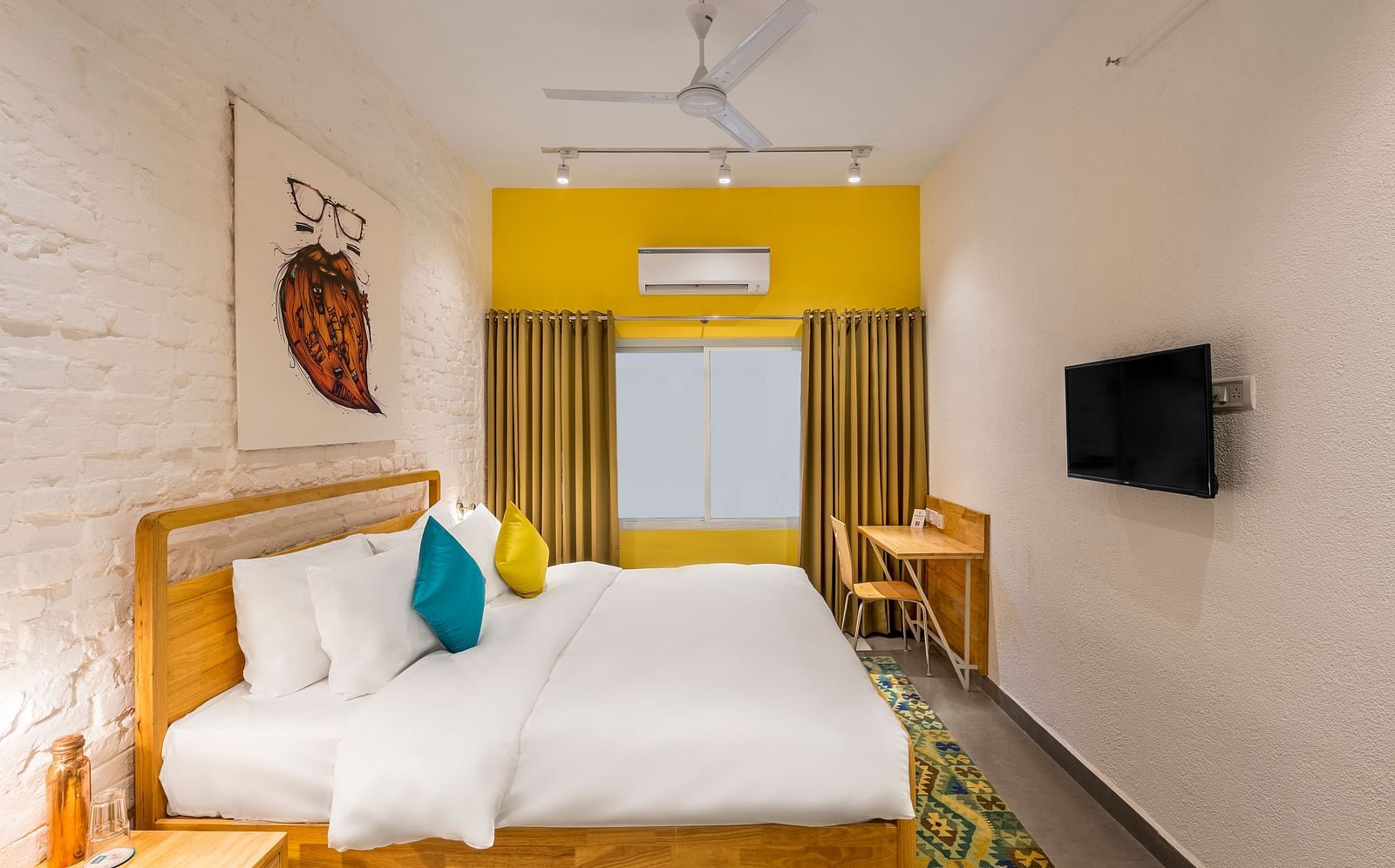 An side angle interior image of the standard room at Bedzzz Varanasi showcases a spacious layout, featuring a comfortable double bed, a cozy floor mat, a stylish study table and chair, a charming coffee table, elegant curtains, an air conditioner for a ref