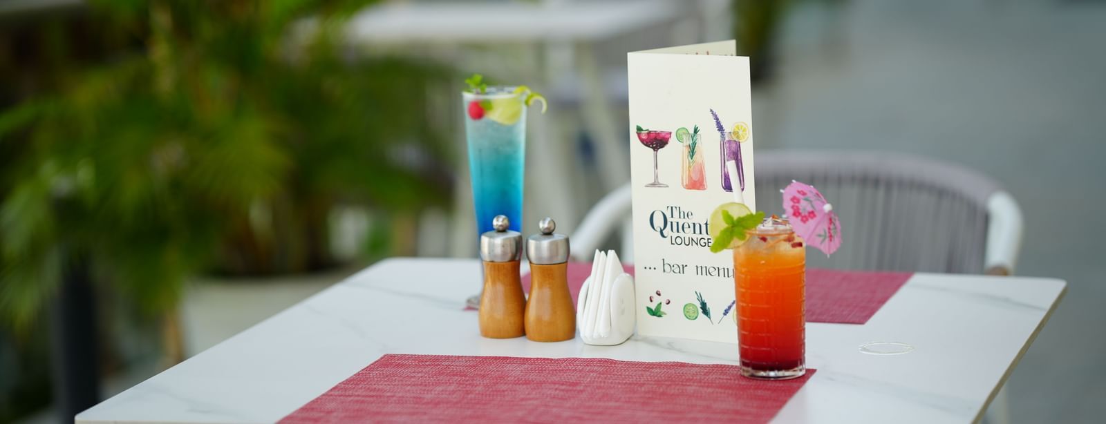 Outdoor dining table with a red drink, a blue drink, menu card and condiments at Grande Bay Resort & Spa.