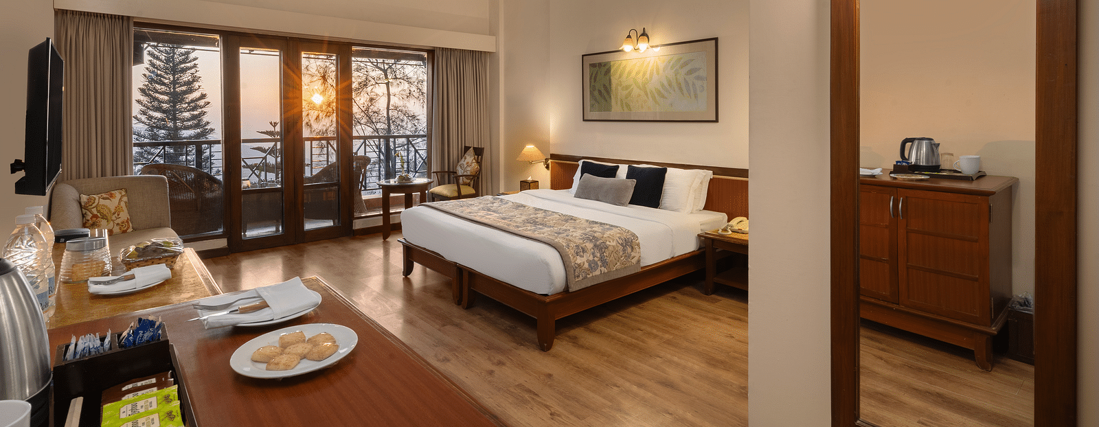 A wide view of a superior room showcasing a bed with side tables, sofas, a large wooden-framed mirror on the wall, and a balcony offering a sunny mountain view  - Baikunth resort, kasauli 