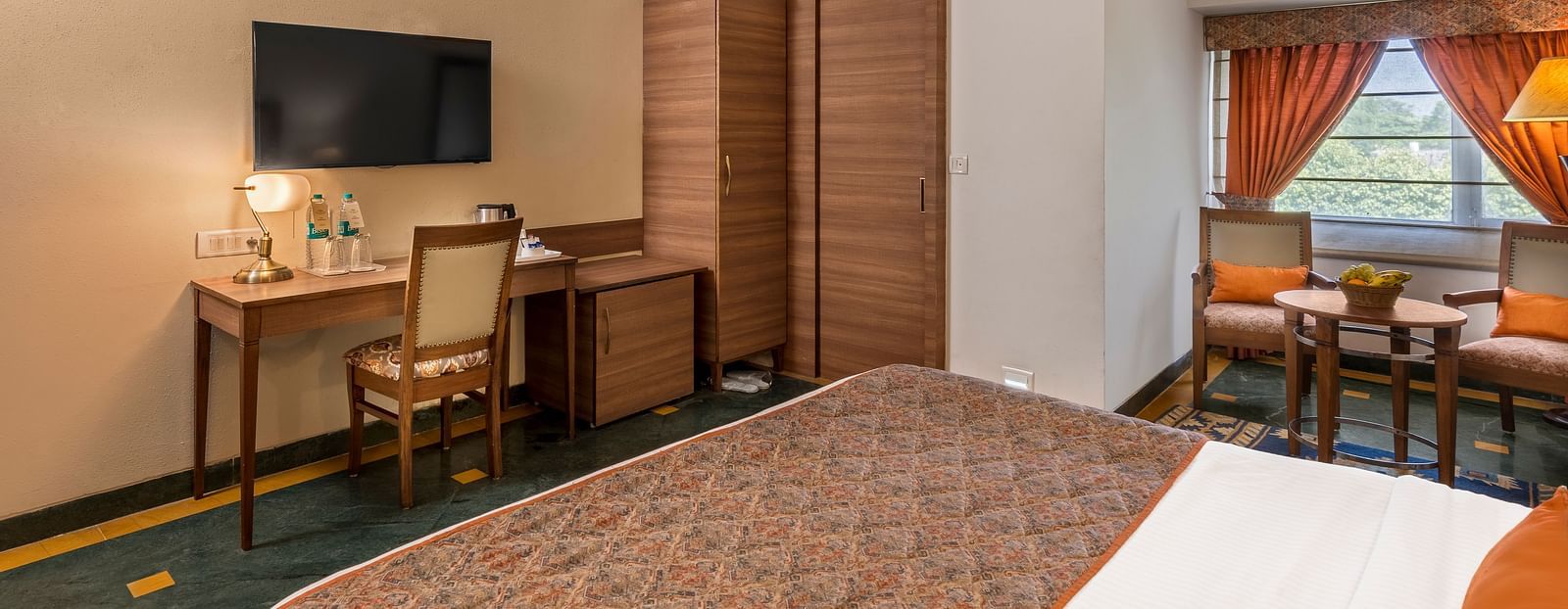 side view of a bedroom with a bed facing a television, below which is a workdesk and chair and a wardrobe beside it - The Manor, Kashipur