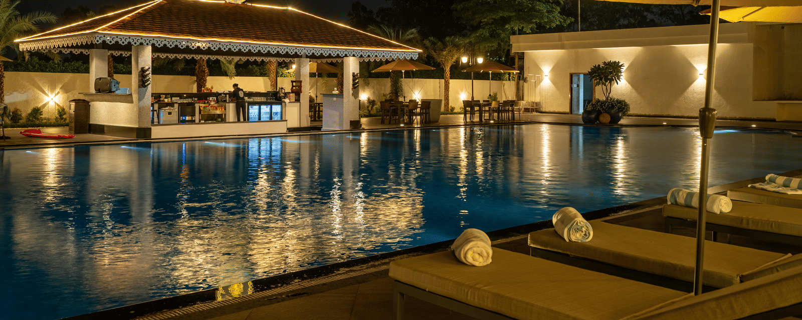 Evening view of the swim-up bar area next to the pool, featuring seating and ambient lighting under an umbrella at Hotel Hukam's Lalit Mahal.