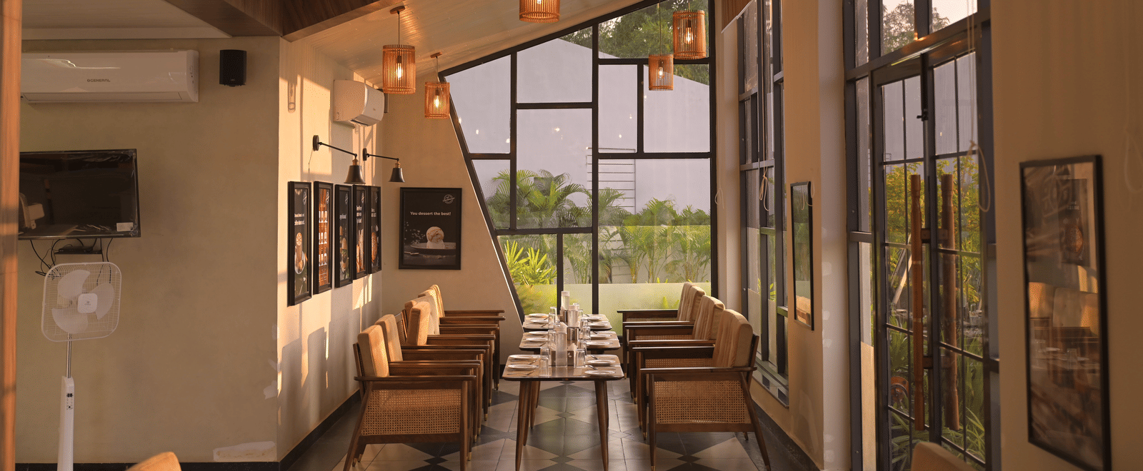 A bright dining corner with hanging lights, stylish decor, and window views of the greenery outside at Hotel Sonar Bangla Mayapur.