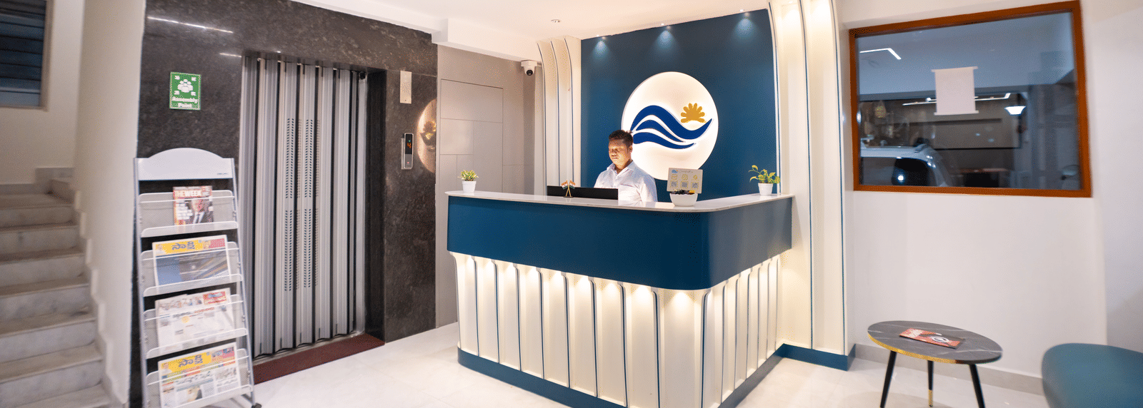 A diagonal view of the reception area with a large desk and a staff member standing behind it, a seating area on one side, and a newspaper stand on the other at Sunrise Business Hotel, Hyderabad.