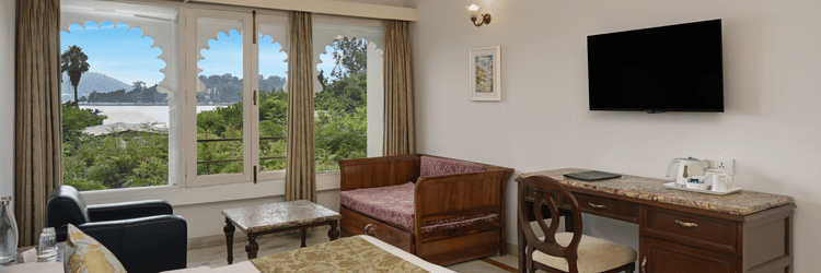 Bedroom at Ram Pratap Palace with a lake view through arched windows, featuring a bed, pillows, two chairs, a coffee table, a daybed, a desk with a chair, a wall-mounted television, an air conditioning unit, and a ceiling fan.