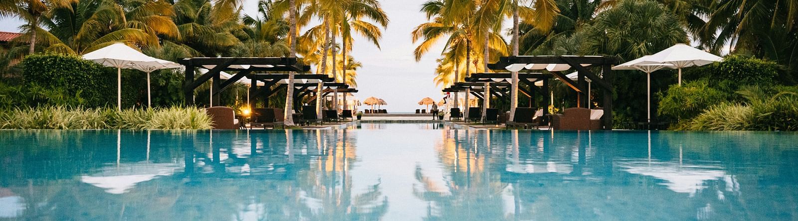 Expansive resort swimming pool reflecting the sky and surrounding palm trees and cabanas.