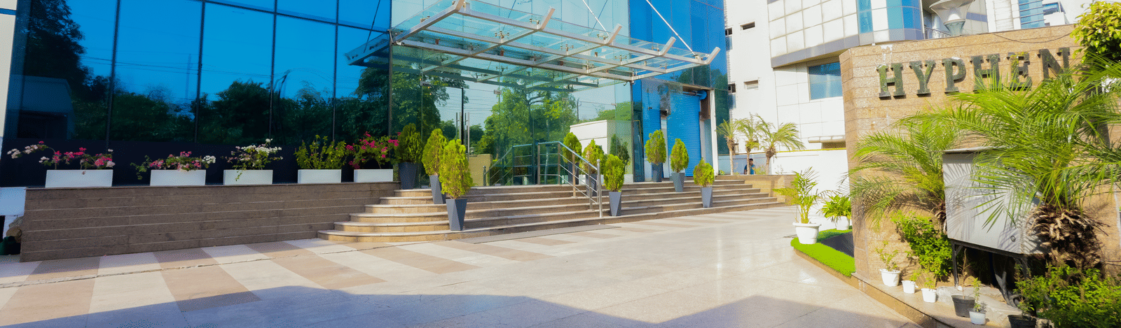 A large, white residential or commercial building with multiple floors stands amidst green landscaping under an overcast sky.