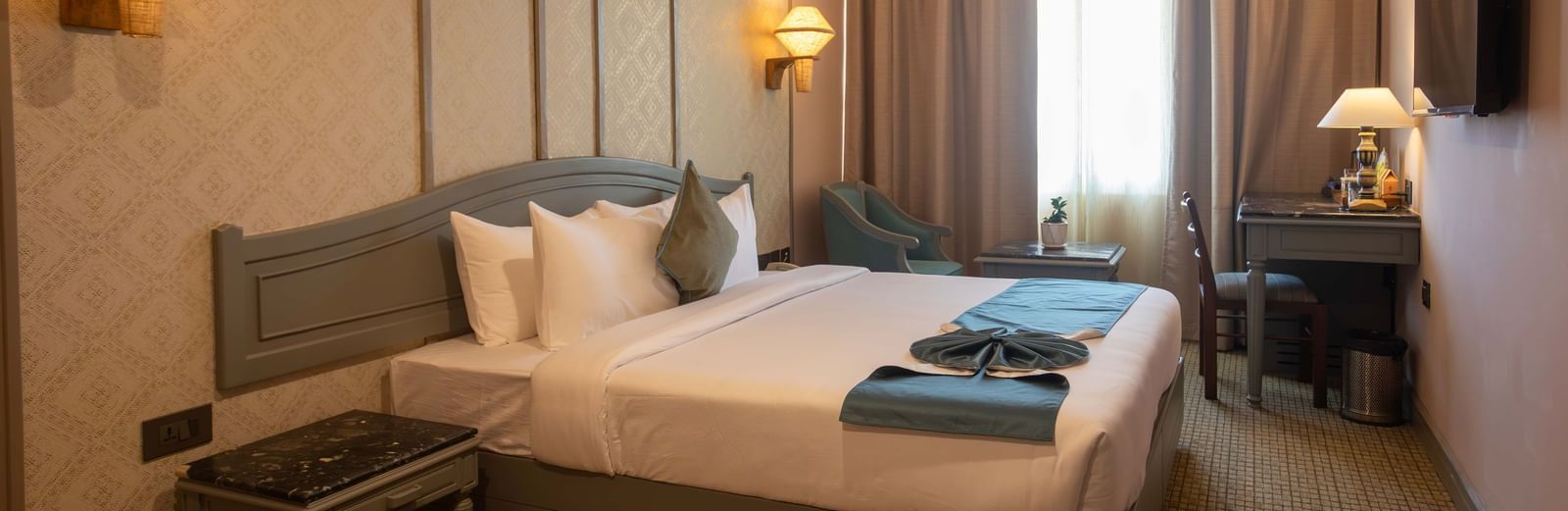 A hotel bedroom with a bed, a bedside table, and curtains covering a window | Shilton Hotels