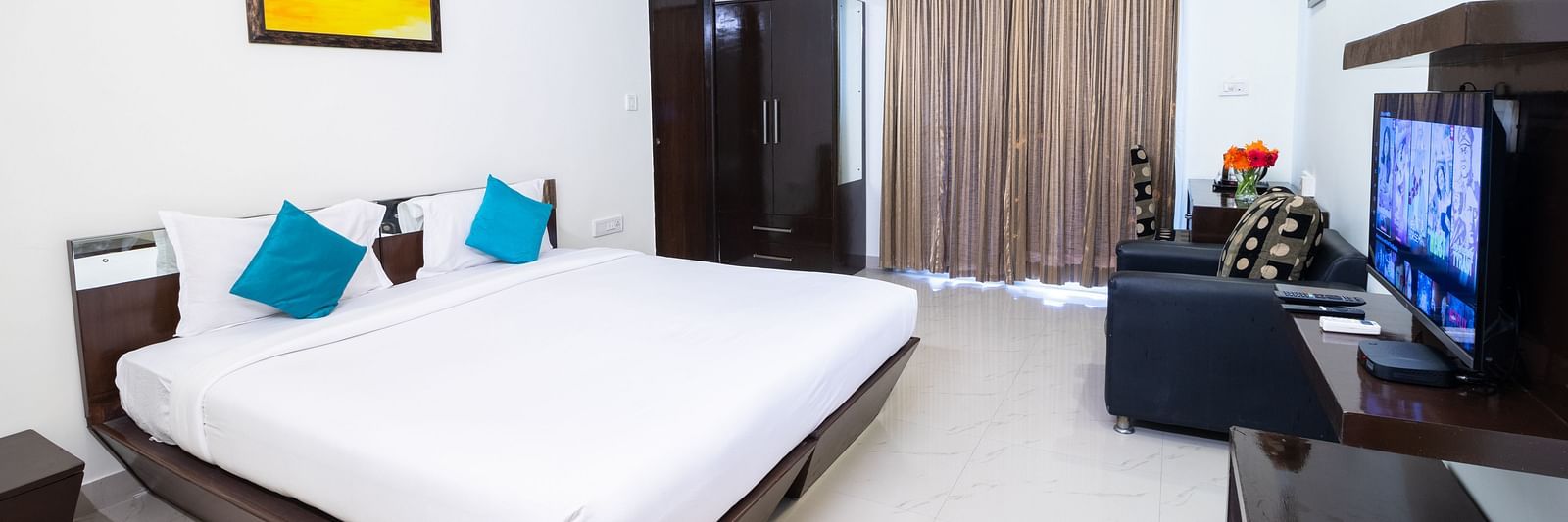 A room offering a double bed, a wall-mounted television, a sofa, and an artwork hung on the wall at Sanctum Suites Domlur, Bangalore.