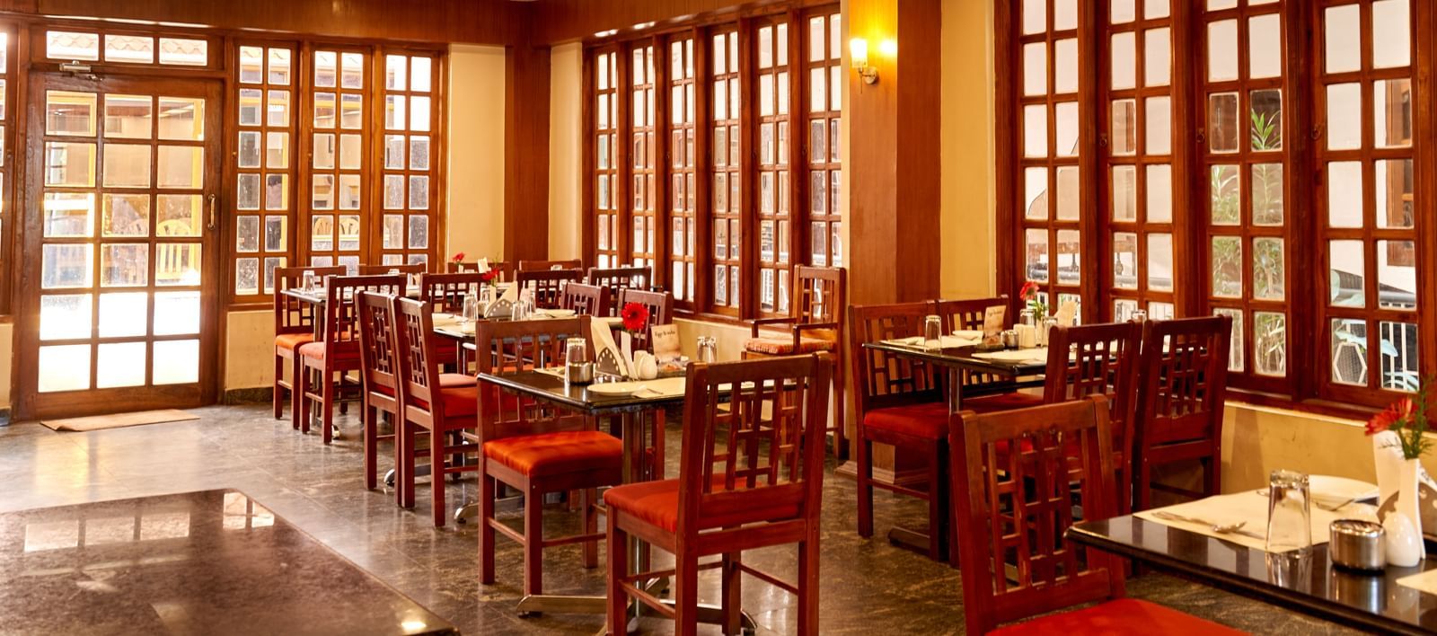 Log shot of the inviting dining area at Hotel Mamallaa Heritage, with red chairs, wooden tables, and natural light filtering through large windows.