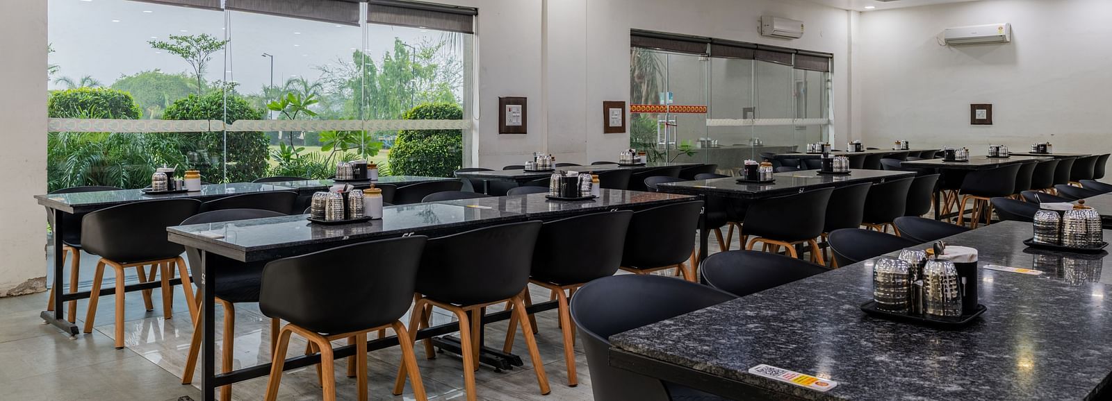 Air-conditioned restaurant of Hotel Highway King, Behror, is furnished with parallel aligned dining tables, water cups and glass windows that offer views of the trees outside at Hotel Highway King, Behror.