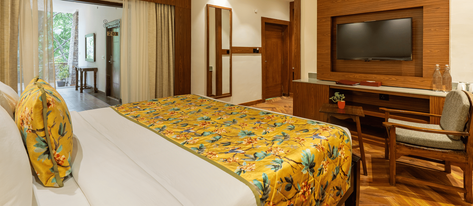 A bedroom featuring a bed, a wooden television unit, a chair and a table, a mirror on a wall, and a verandah with trees outside | Summit by the Ganges Beach Resort & Spa, Rishikesh