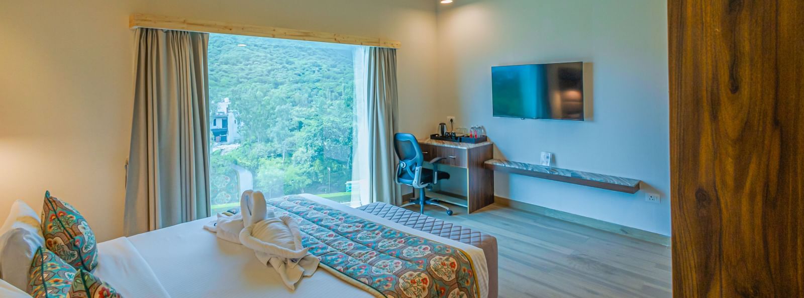 sajjan villa room at Bamboo Saa Resort and Spa, Udaipur featuring a bed, a flat TV screen, a window with curtains, a ceiling fan and a  work desk and chair.