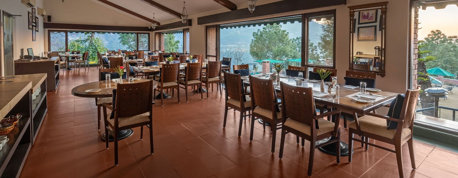 A restaurant with glass windows offering a beautiful mountain view, featuring dining tables arranged with chairs, a mirror on the wall, and stylish pendant lights overhead -  Baikunth resort, kasauli 