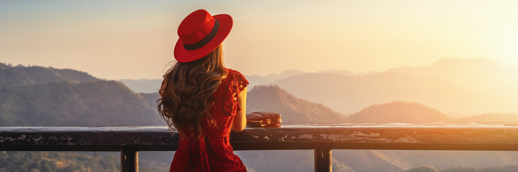 Person in a red outfit and hat sits on a stool at a scenic overlook, gazing at layered mountains during sunrise or sunset, with a drink placed on the counter in front.