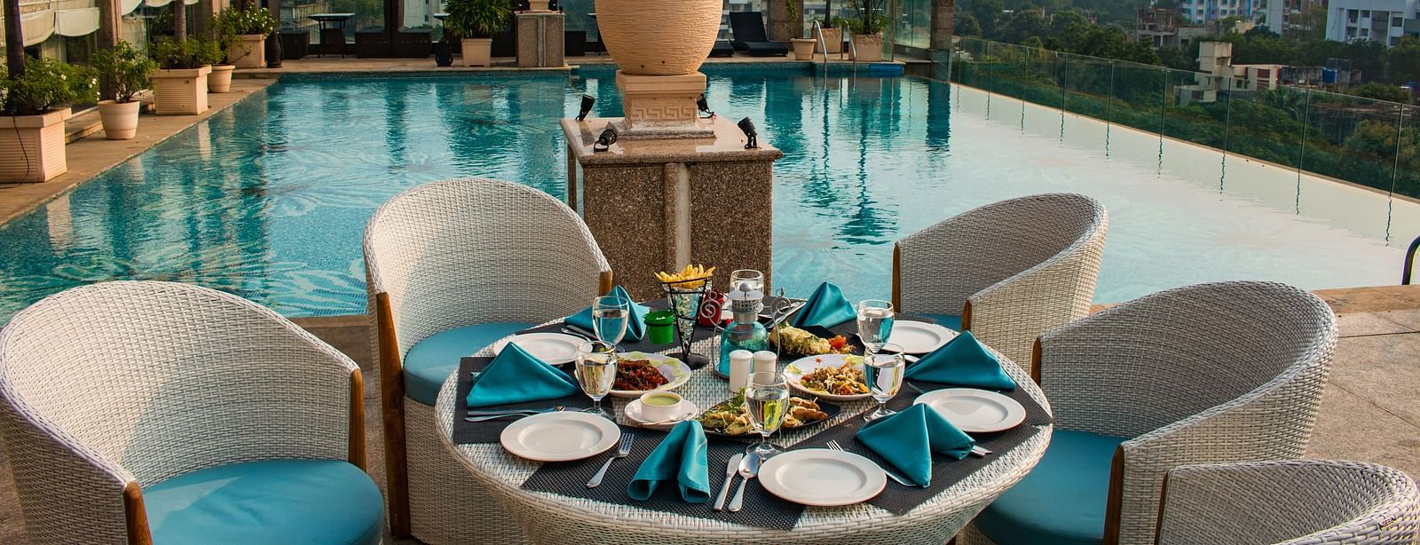 An outdoor dining at Hotel Ashok Nagpur featuring a round table with place settings for four, surrounded by cushioned chairs, and a swimming pool and cityscape in the background. 