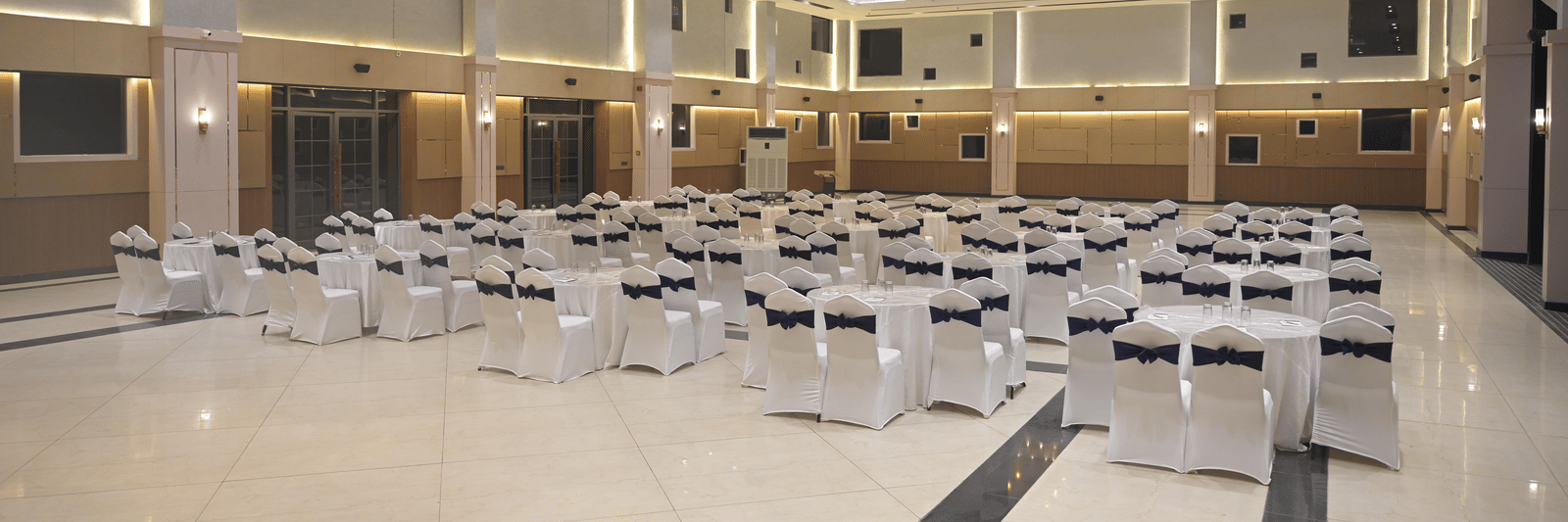 A panoramic view of the banquet hall arranged for a gathering, featuring bright chandeliers and neatly placed round tables at Hotel Sonar Bangla Mayapur.