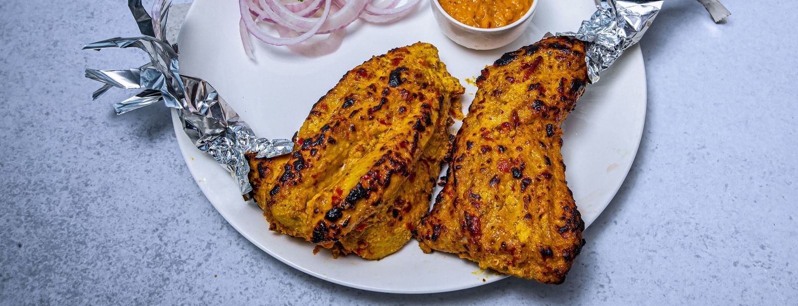 A plate of grilled tandoori chicken legs wrapped in foil at the ends, served with sliced onions, a lemon wedge on a white background at The Hamuse Hotel, Kodaikanal.