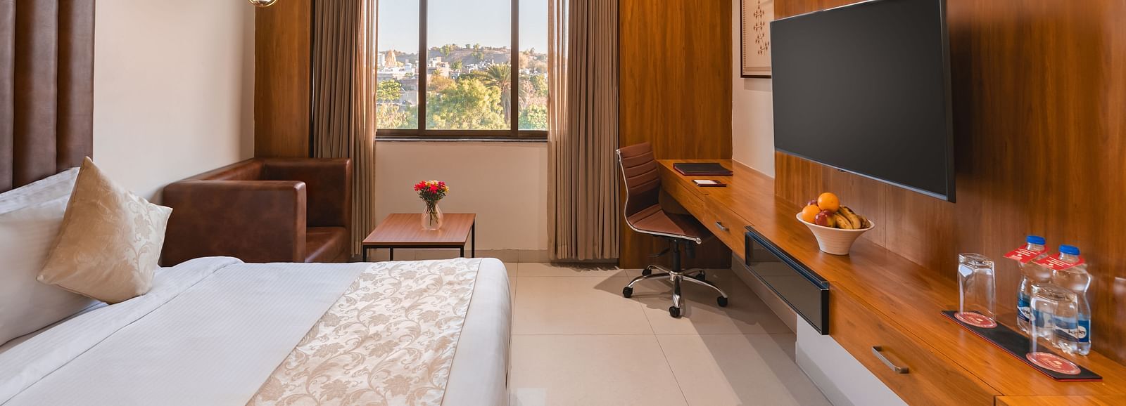 A well-furnished hotel room includes a comfortable bed, a seating area, a desk with a chair, and a mounted television - Lords Eco Inn Ambaji.