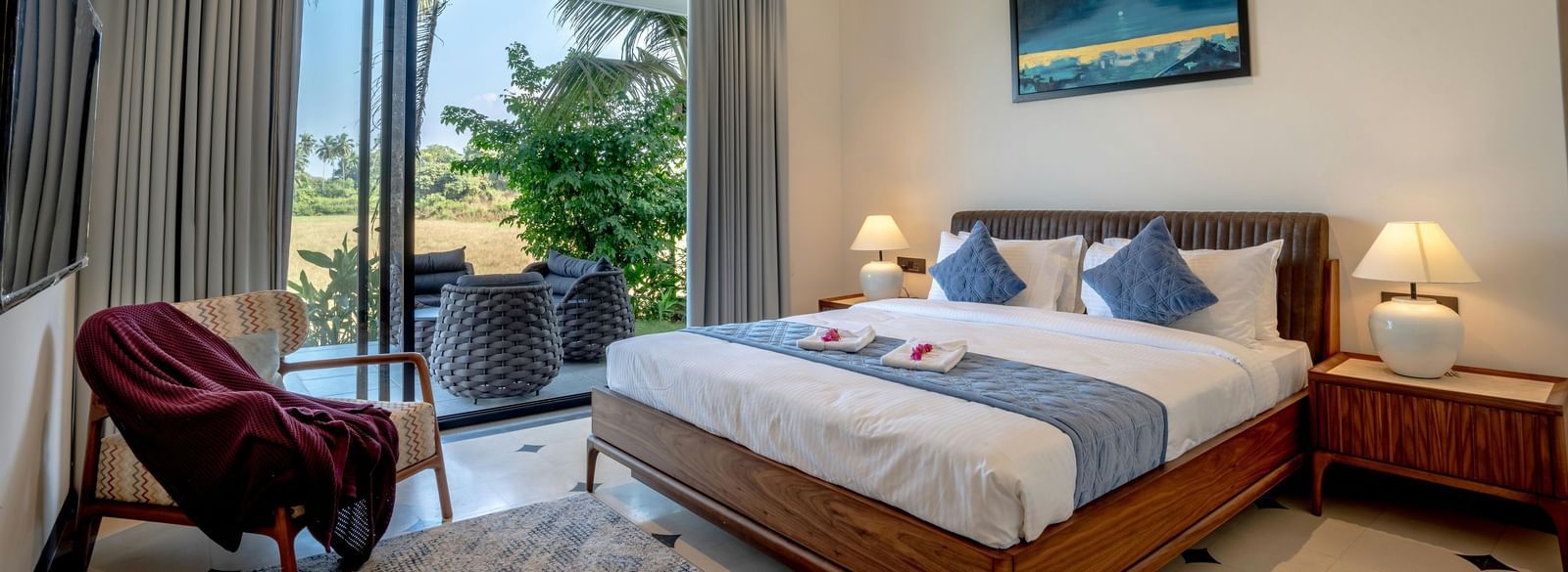 A room with balcony, spacious bed with bedside tables and lamps, artwork above the headboard, and outdoor view through glass doors