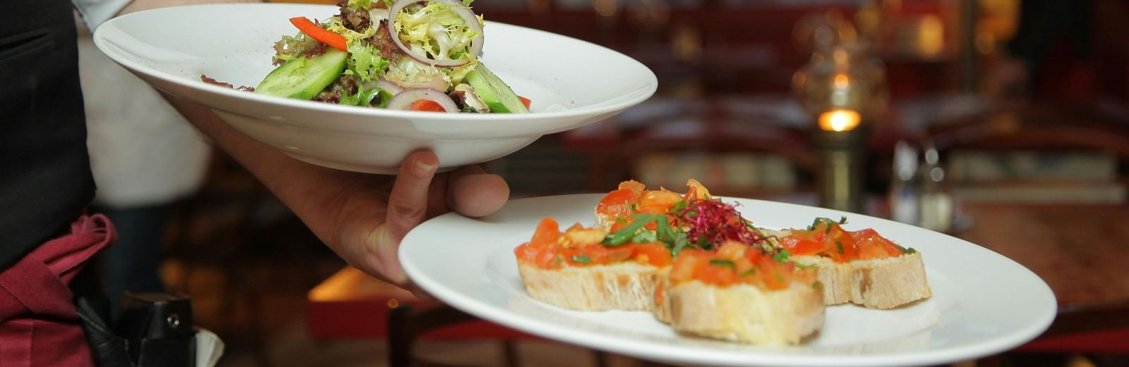 A waiter in a white shirt and black vest carefully carries a plate of salad and a plate of bruschetta in a warmly lit restaurant setting.