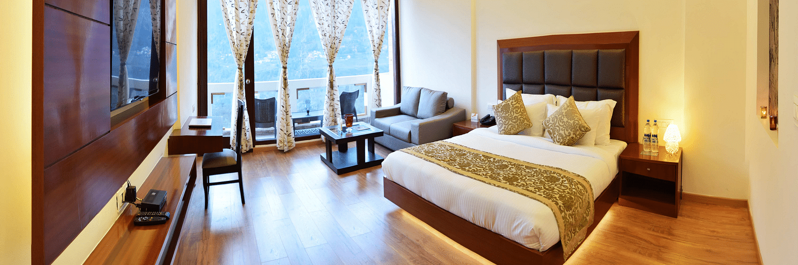 The image shows a spacious and luxurious hotel room with a king size bed, a sitting area with a sofa and coffee table, and a large window with a view.  | DLS Serenity Resort and Spa
